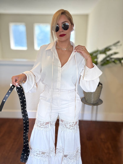 White long-sleeve jumpsuit with lace panels, deep V neckline, and black studded belt
